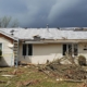 Storm-damaged Minnesota home with missing shingles, broken siding, and debris in the yard as repairs begin